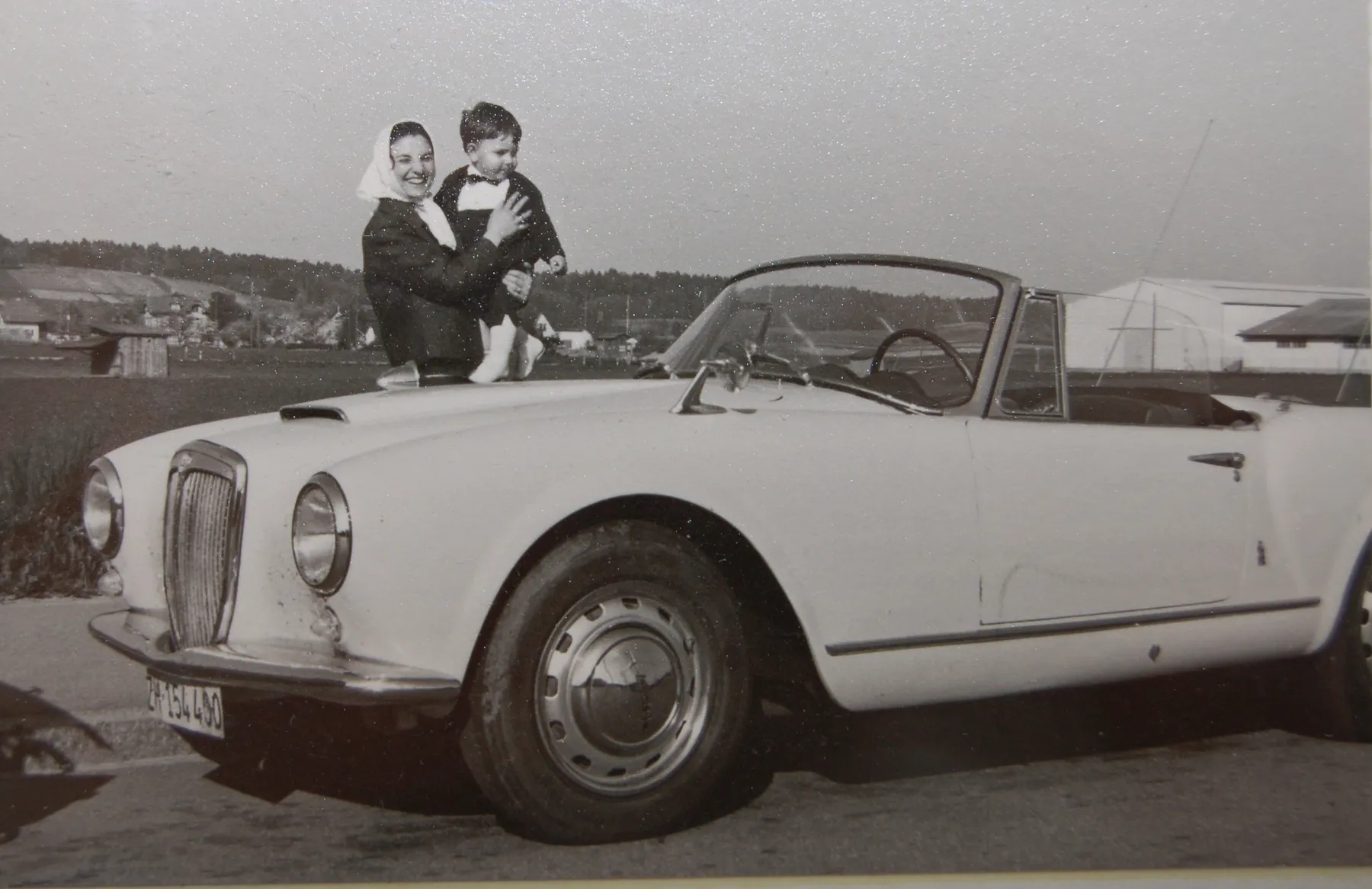 Historische Schwarz-Weiss-Aufnahme: Mutter mit Kind auf dem Arm neben einem offenen Lancia-Cabriolet